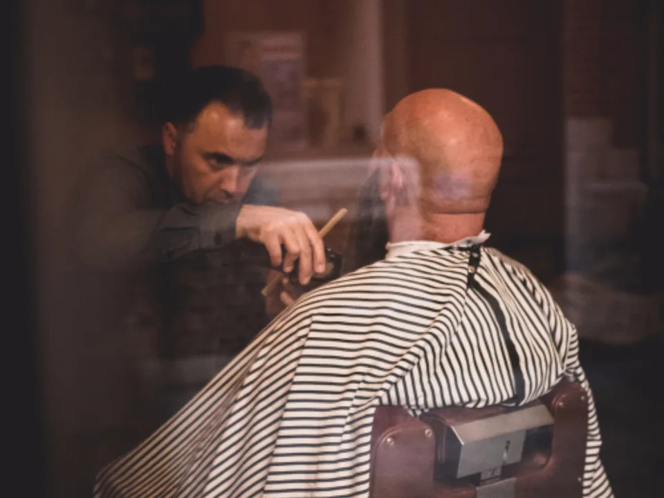 Seen through the shop window: a barber part-way through a cut, the client under a striped cape.