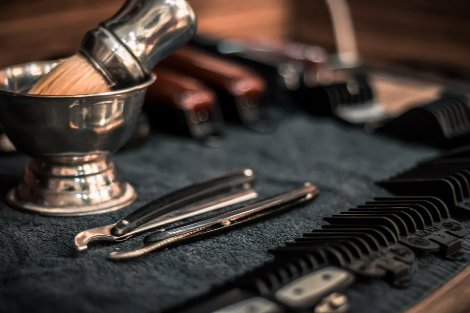 A barber's kit laid out on black — brass lather bowl, badger brush, folded straight razors and combs.